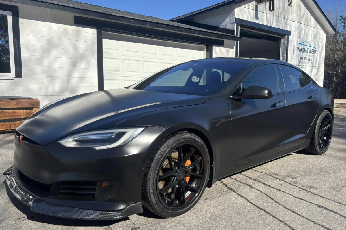 A car with matte black wrap and tinted windows, parked in front of a garage.